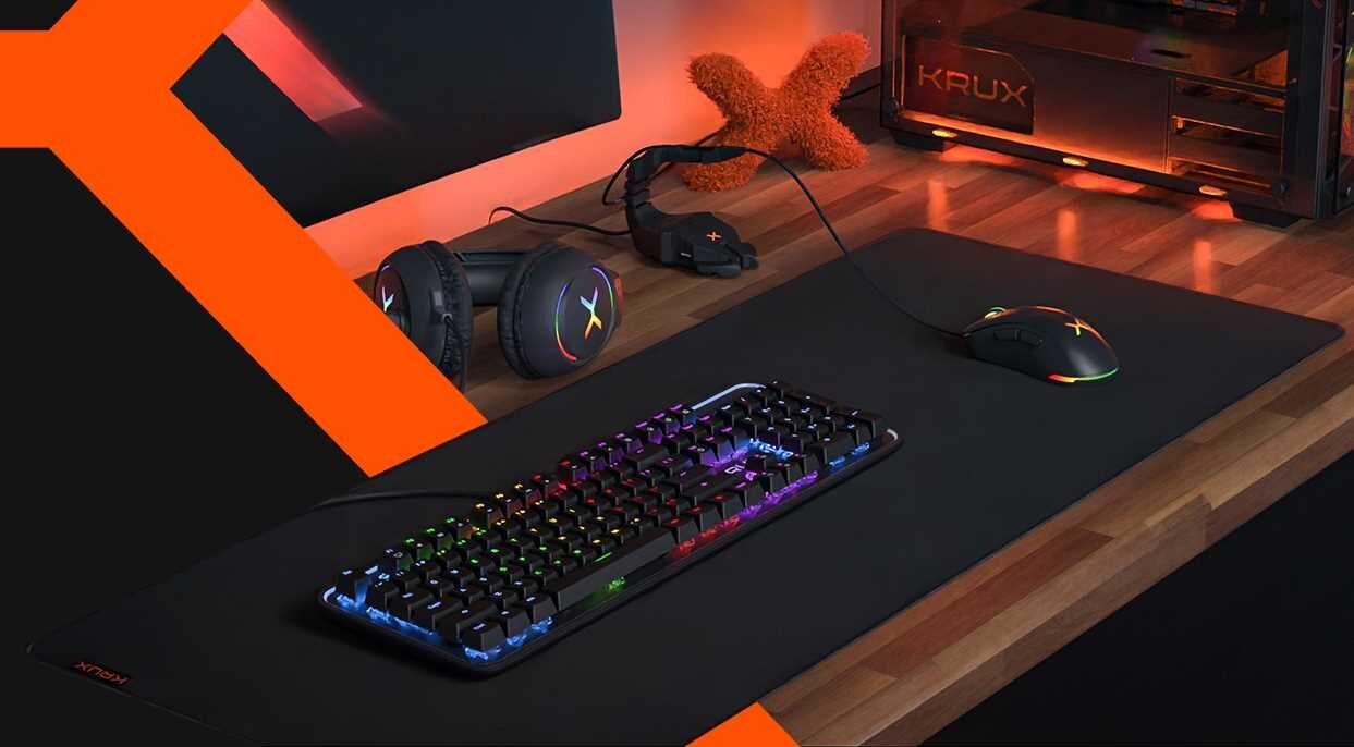 Na drewnianym biurku znajduje się duża podkładka, na której umieszczono klawiaturę z podświetleniem RGB oraz myszkę. W tle widoczne są słuchawki i komputer z logo \'KRUX\'.