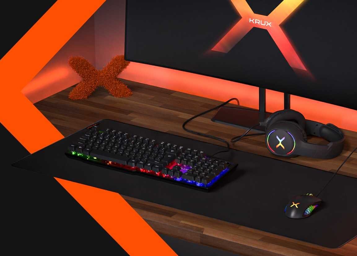 Na drewnianym biurku znajduje się klawiatura z podświetleniem RGB, mysz oraz słuchawki z logo \'X\'. W tle widoczny jest monitor z logo \'KRUX\' oraz dekoracja w kształcie litery \'X\'.