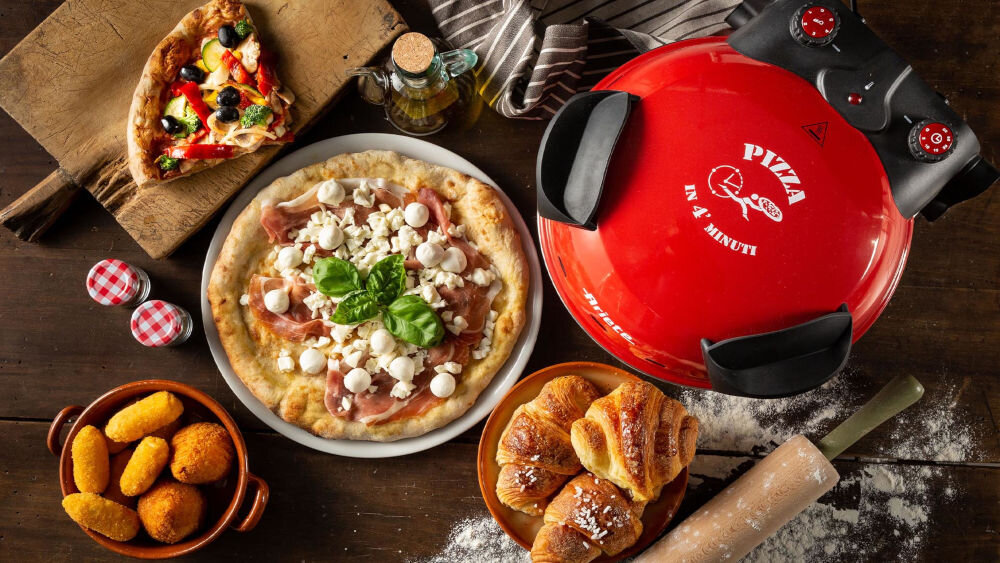 Na stole znajduje się czerwony piec do pizzy z napisem \'PIZZA IN 4 MINUTI\', obok którego leży gotowa pizza z mozzarellą i bazylią. Wokół pieca widoczne są różne potrawy, w tym kawałek pizzy, croissanty i panierowane przekąski.