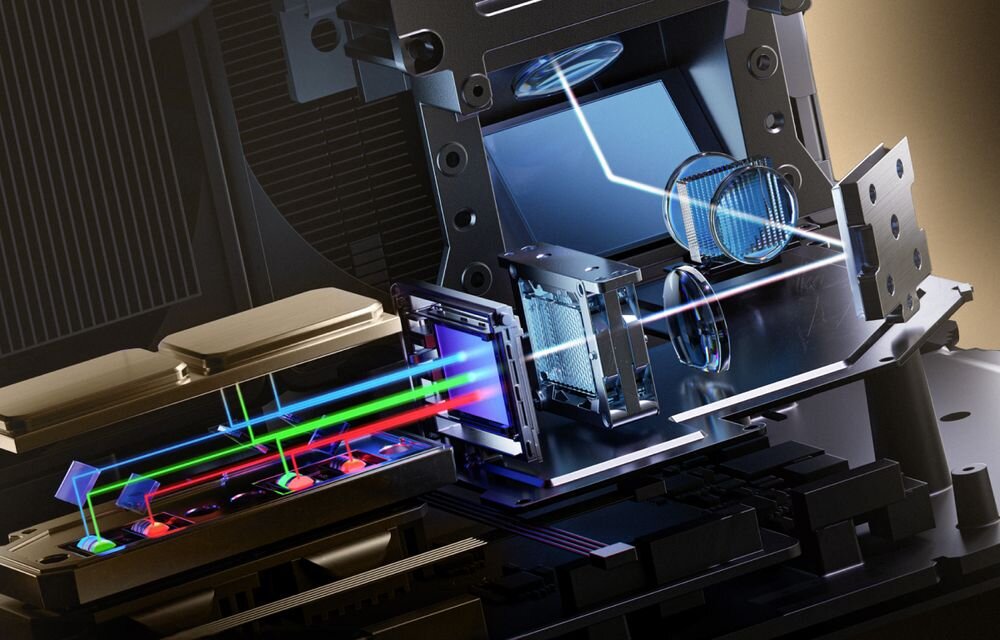 Obraz przedstawia wnętrze projektora laserowego z widocznymi elementami optycznymi i kolorowymi wiązkami światła. Widać soczewki, pryzmaty i inne komponenty technologiczne.