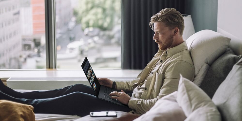 Mężczyzna w beżowej koszuli pracujący na laptopie ASUS ProArt P16 leżąc na łóżku w jasnym pokoju hotelowym z widokiem na miasto przez okno - praca kreatywna 