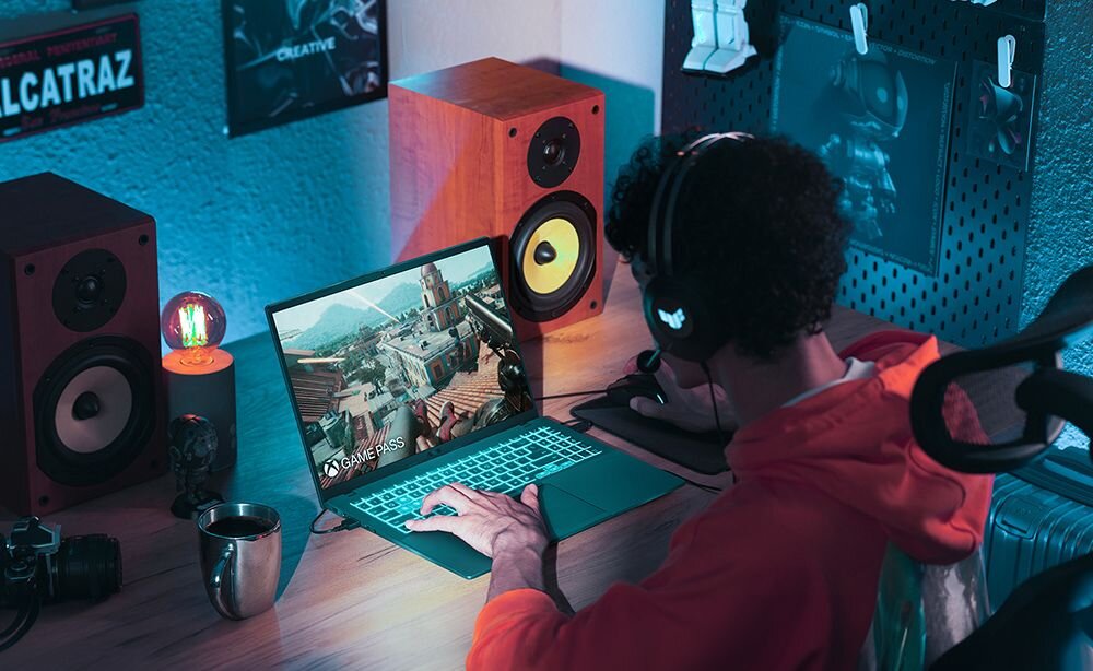 Gracz w czerwonej bluzie korzystający z laptopa ASUS V16 podczas rozgrywki ze słuchawkami głośnikami drewnianymi aparatem i kubkiem na biurku - Procesor Intel Core Ekran 16 cali MIL-STD-810H 