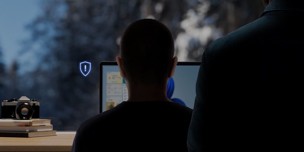 Dwóch mężczyzn przy biurku z laptopem ASUS ZenBook wyposażonym w kamerę AI z niebieską ikoną tarczy bezpieczeństwa oraz aparatem analogowym i książkami w tle - automatycznie ściemnia ekran po odwróceniu wzroku