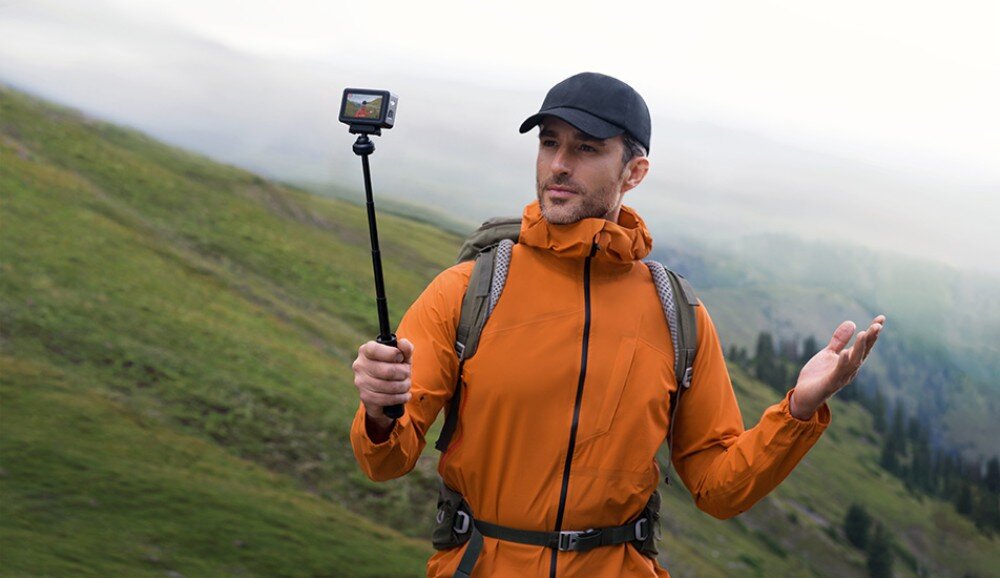 Kamera sportowa DJI Osmo Action 6 Mężczyzna w pomarańczowej kurtce trzymający kamerę na wysięgniku w górskim otoczeniu, obsługa, ustawienia, przenoszenie nagrań