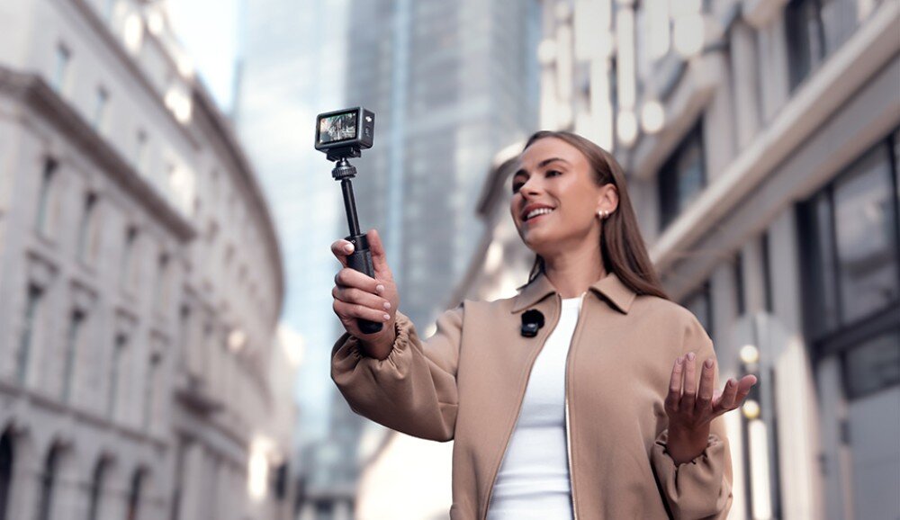 Kamera sportowa DJI Osmo Action 6 Kobieta trzymająca kamerę na uchwycie i mówiąca do obiektywu na tle budynków, dźwięk, mikrofony, redukcja szumów