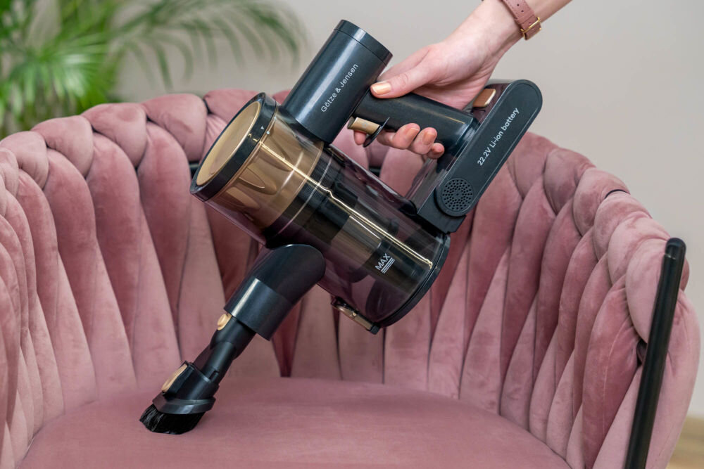 Osoba trzyma odkurzacz ręczny GOTZE & JENSEN VV300 nad różowym, welurowym fotelem. Na odkurzaczu widoczne są napisy: \'GOTZE & JENSEN\' oraz \'22.2V Li-ion battery\'.