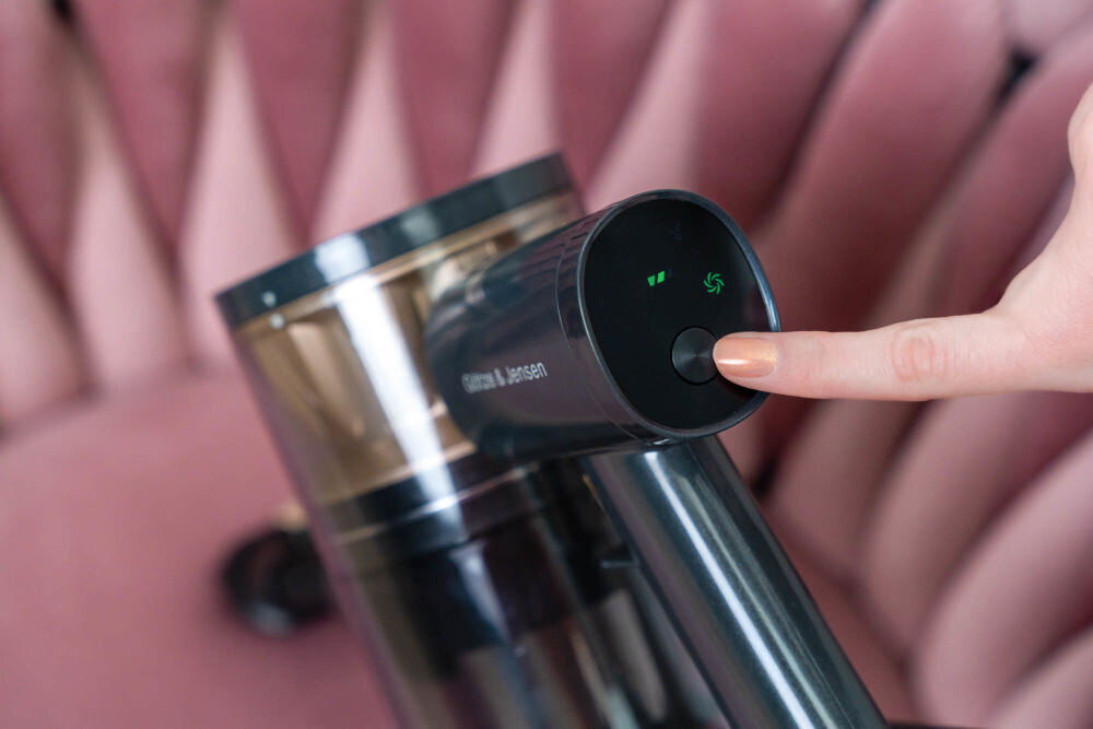 Osoba naciska przycisk na odkurzaczu z widocznym logo \'Gotze & Jensen\'. W tle znajduje się różowa tapicerka.