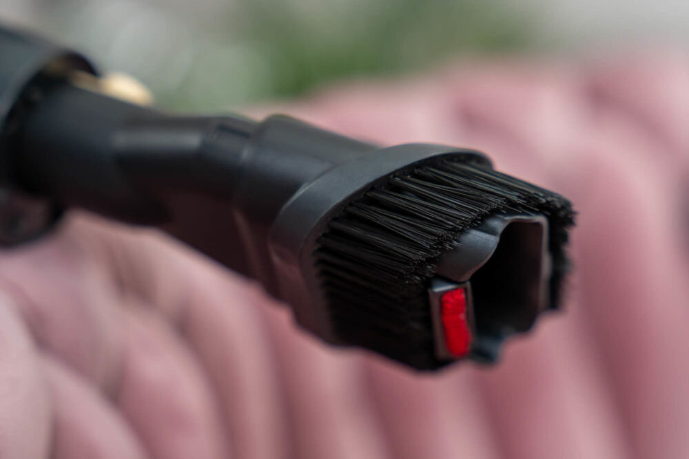 Zbliżenie na końcówkę odkurzacza z czarnymi włosiami i czerwonym elementem. Tło jest rozmyte, w odcieniach różu.