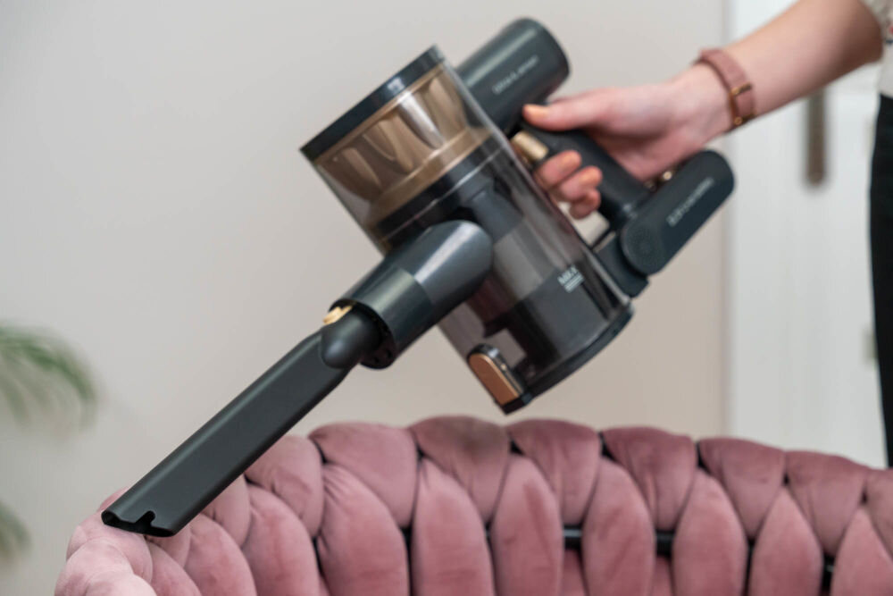 Osoba trzyma odkurzacz ręczny z widocznym logo \'GOTZE & JENSEN\'. Odkurzacz jest używany do czyszczenia różowego, pikowanego fotela.