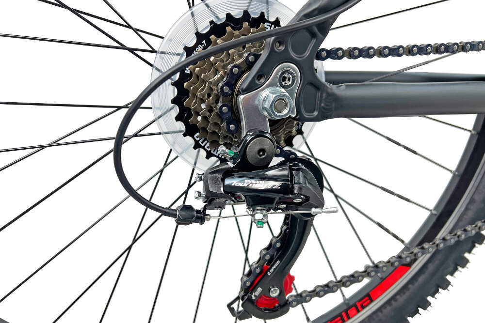 Zbliżenie na tylną przerzutkę roweru z widocznym mechanizmem zębatek i łańcuchem. Na przerzutce widoczny jest napis \'Shimano Tourney TZ\'.