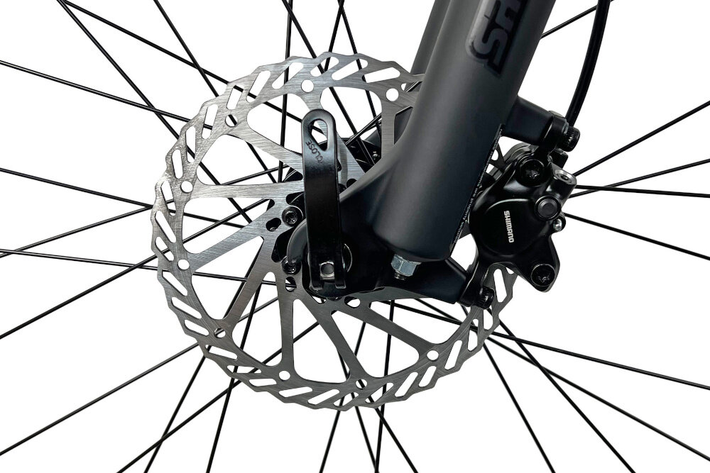 Zbliżenie na tarczę hamulcową roweru z widocznym zaciskiem hamulcowym marki Shimano. W tle widać szprychy koła.