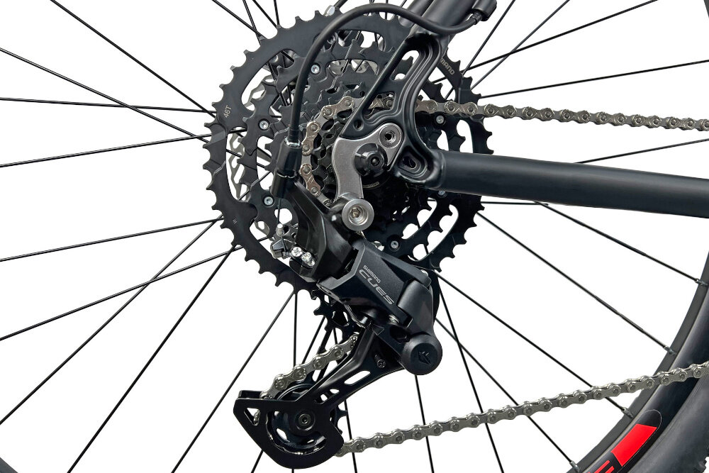 Zdjęcie przedstawia mechanizm przerzutki rowerowej z widocznym łańcuchem i zębatkami. Elementy są w kolorze czarnym, a część ramy roweru jest również widoczna.