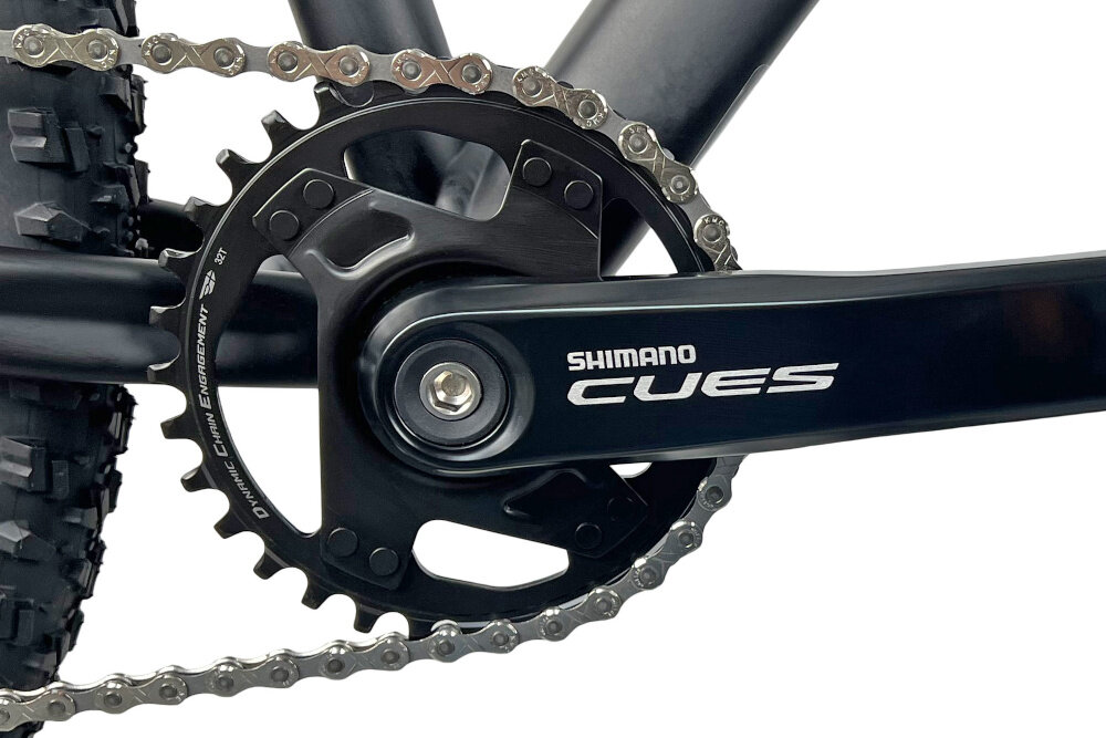 Zbliżenie na mechanizm korbowy roweru z widocznym napisem \'SHIMANO CUES\' na ramieniu korby. Łańcuch jest nałożony na zębatkę, a opona ma wyraźny bieżnik.