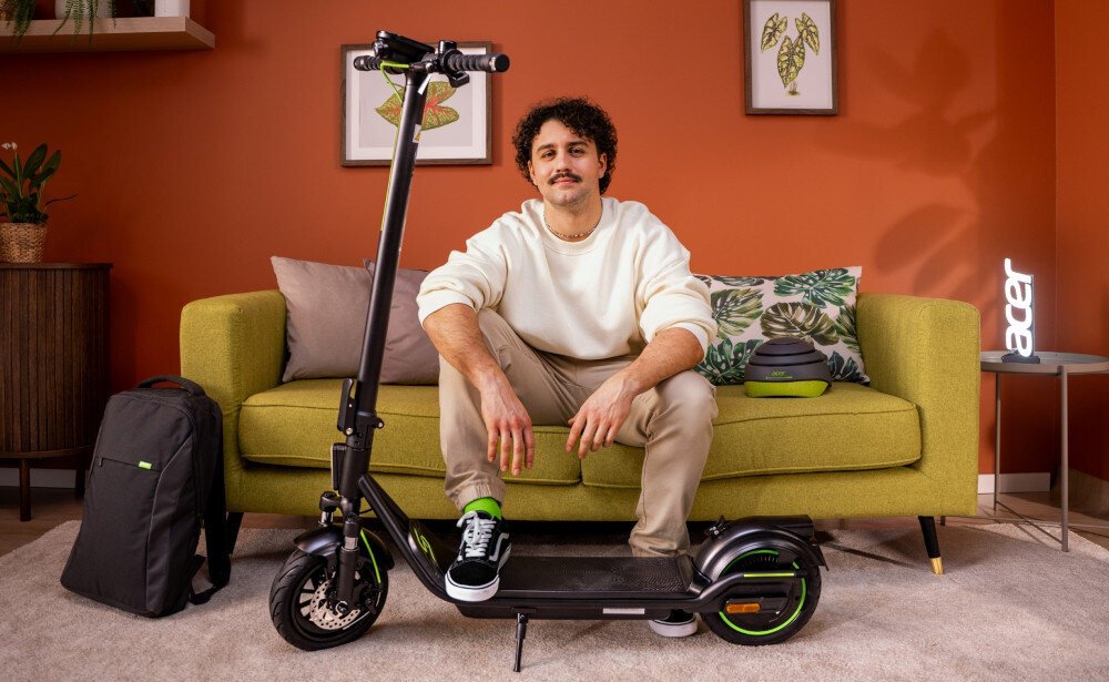 Hulajnoga elektryczna ACER ES Series 5 Select 60km 500W Amortyzacja 10 cali Czarny pokój pomarańczowa ściana żółta kanapa osoba siedzi na kanapie przed osobą stoi hulajnoga stabilność wygoda środek transportu jazda po mieście