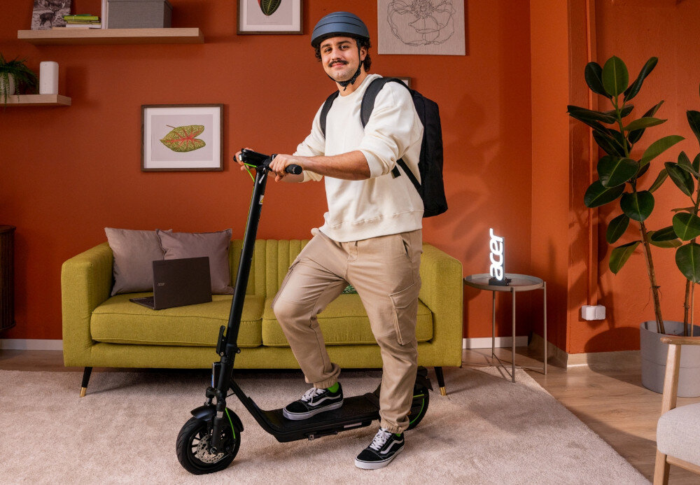 Hulajnoga elektryczna ACER ES Series 5 Select 60km 500W Amortyzacja 10 cali Czarny pokój pomarańczowa ściana żółta kanapa osoba stoi na podłodze jedną stopę opiera o podest hulajnogi drugą o podłogę sprężynowe zawieszenie wytrzymałość i skuteczność amortyzacja bardzo dobrze tłumi drgania oraz wstrząsy poprawia wygodę stabilność i bezpieczeństwo