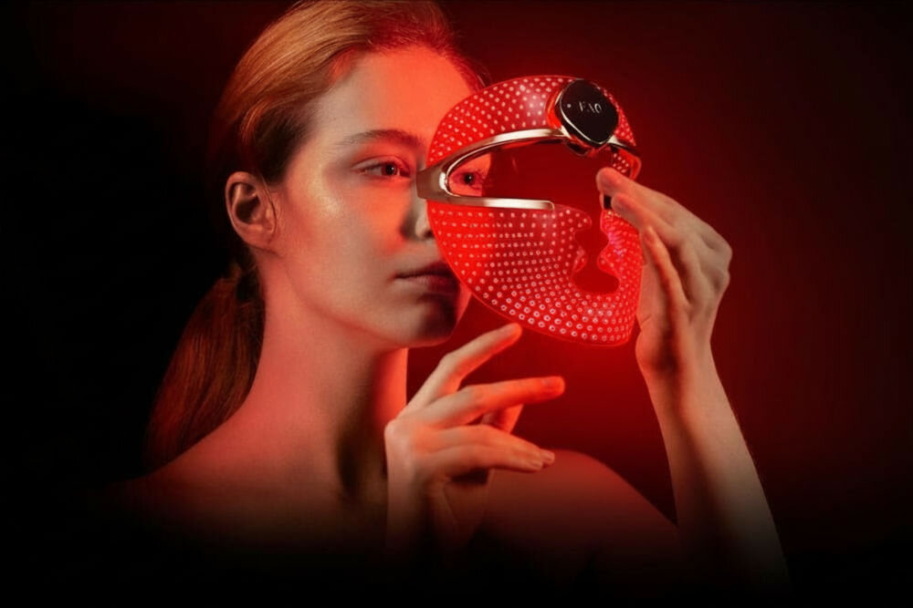Maska LED FOREO Faq 202 Kobieta o jasnej karnacji w ciemnym otoczeniu trzyma w dłoniach czerwoną maskę LED na twarz, która jest włączona i świeci
 
