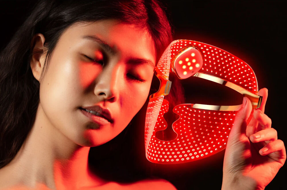 Maska LED FOREO Faq 202 Zbliżenie na kobietę w czerwonym świetle, z zamkniętymi oczami, trzymającą w ręku świecącą na czerwono maskę LED z widocznymi punktami świetlnymi.
 