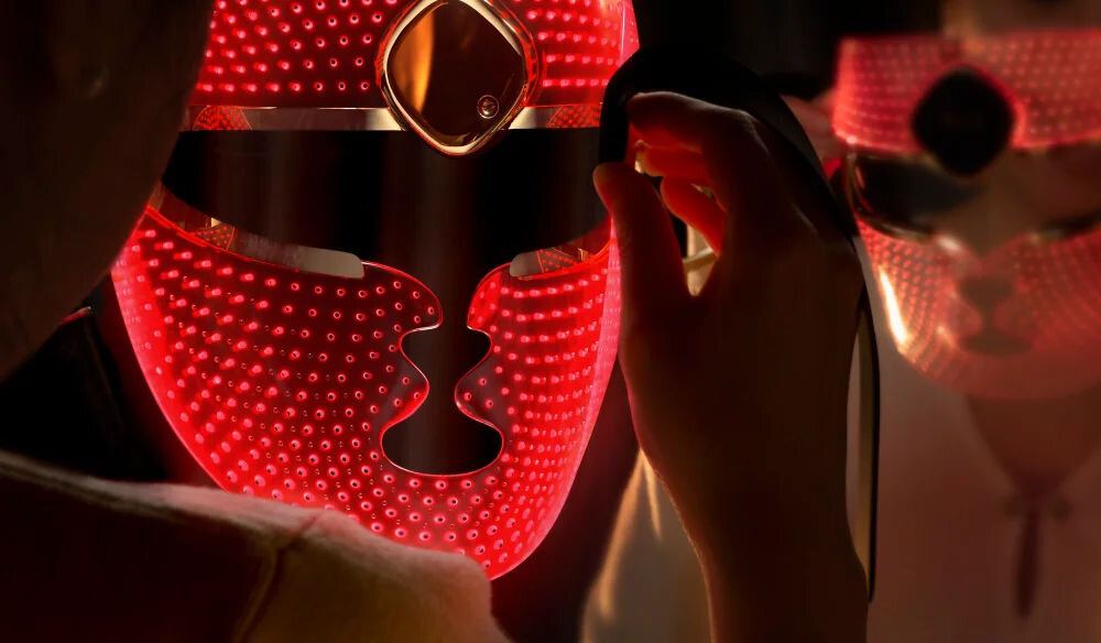 Maska LED FOREO Faq 202 Zbliżenie na detale świecącej na czerwono maski LED na twarz, z wyraźnie widoczną liczną perforacją i punktami świetlnymi oraz elementami w kolorze różowego złota.
 