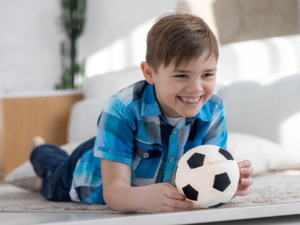 Lampka nocna INNOGIO GIO Piłka GIO-155 Uśmiechnięty chłopiec leży na dywanie i trzyma w dłoniach świecącą lampkę nocną w kształcie czarno-białej piłki nożnej marki innoGIO.