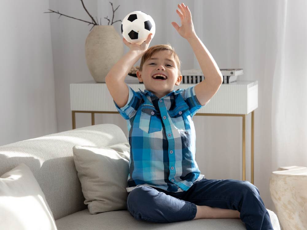 Lampka nocna INNOGIO GIO Piłka GIO-155 Radosny chłopiec siedzi na jasnej kanapie i unosi w górę silikonową lampkę nocną przypominającą piłkę nożną.