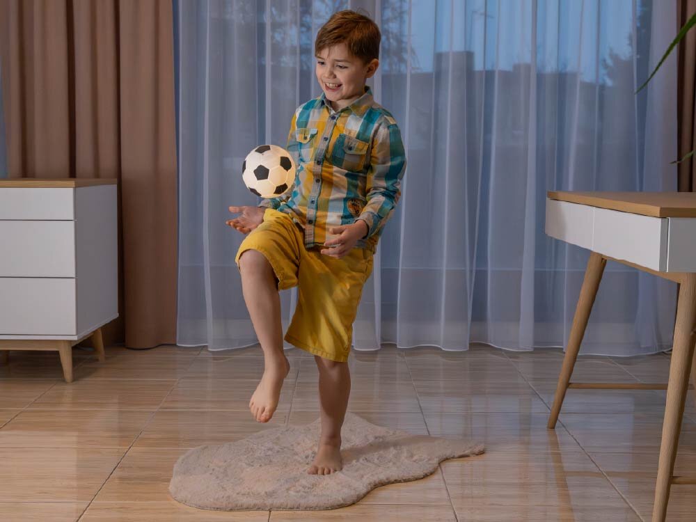 Lampka nocna INNOGIO GIO Piłka GIO-155 Chłopiec w pokoju dziecięcym bawi się świecącą lampką w kształcie piłki, podrzucając ją kolanem niczym prawdziwą piłkę nożną.