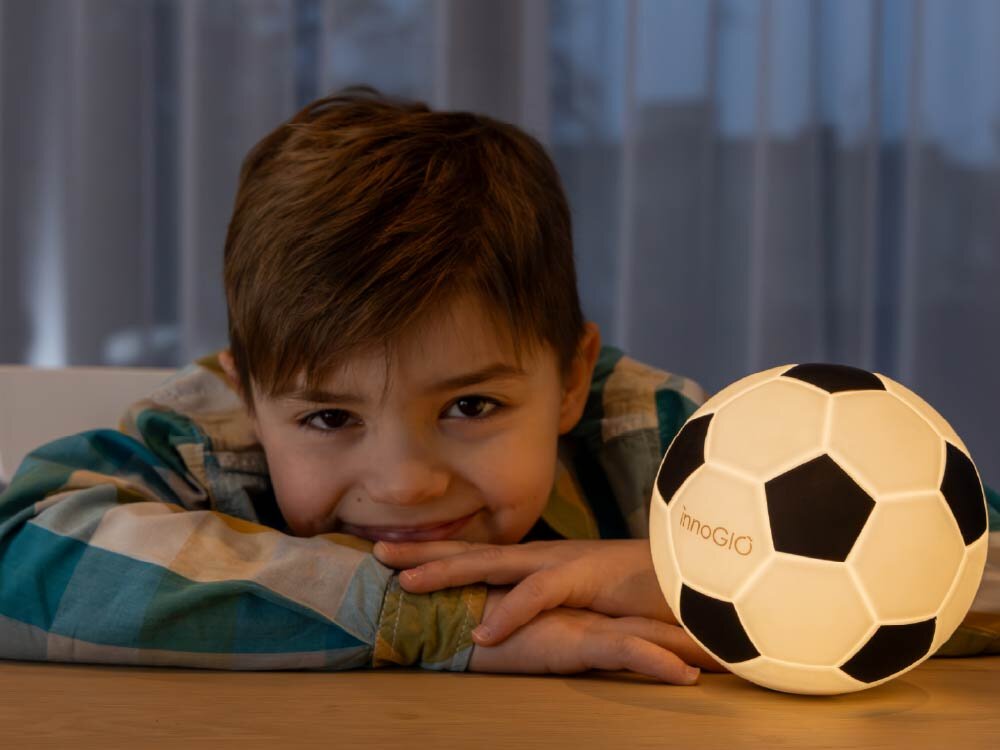 Lampka nocna INNOGIO GIO Piłka GIO-155 Chłopiec opiera głowę na dłoniach na biurku obok świecącej lampki nocnej innoGIO w kształcie piłki nożnej.