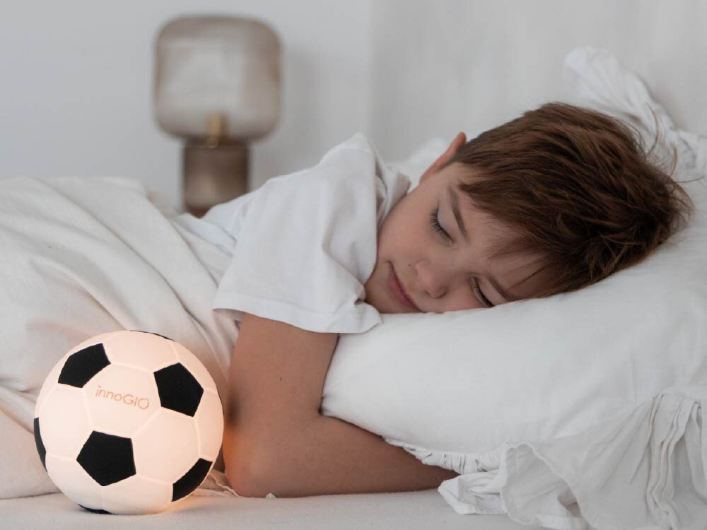 Lampka nocna INNOGIO GIO Piłka GIO-155 Dziecko śpi spokojnie w białej pościeli, a obok niego na łóżku świeci delikatnym światłem lampka nocna w formie piłki nożnej.