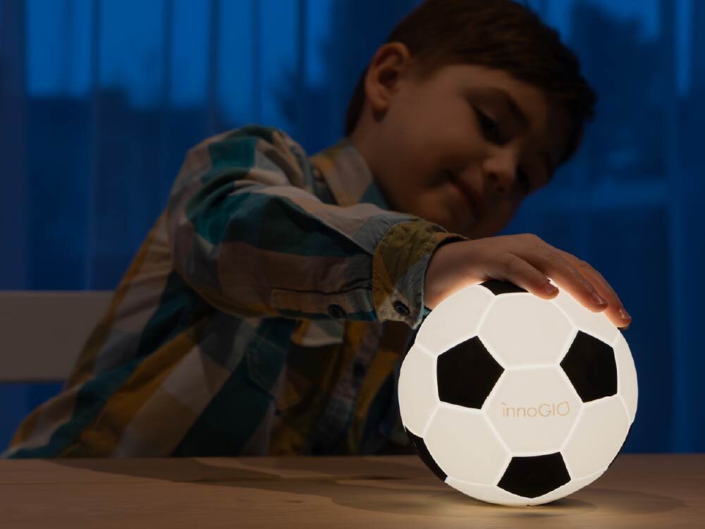 Lampka nocna INNOGIO GIO Piłka GIO-155 Chłopiec w przyciemnionym pokoju kładzie dłoń na świecącej lampce nocnej w kształcie piłki, demonstrując jej miękką, silikonową obudowę.