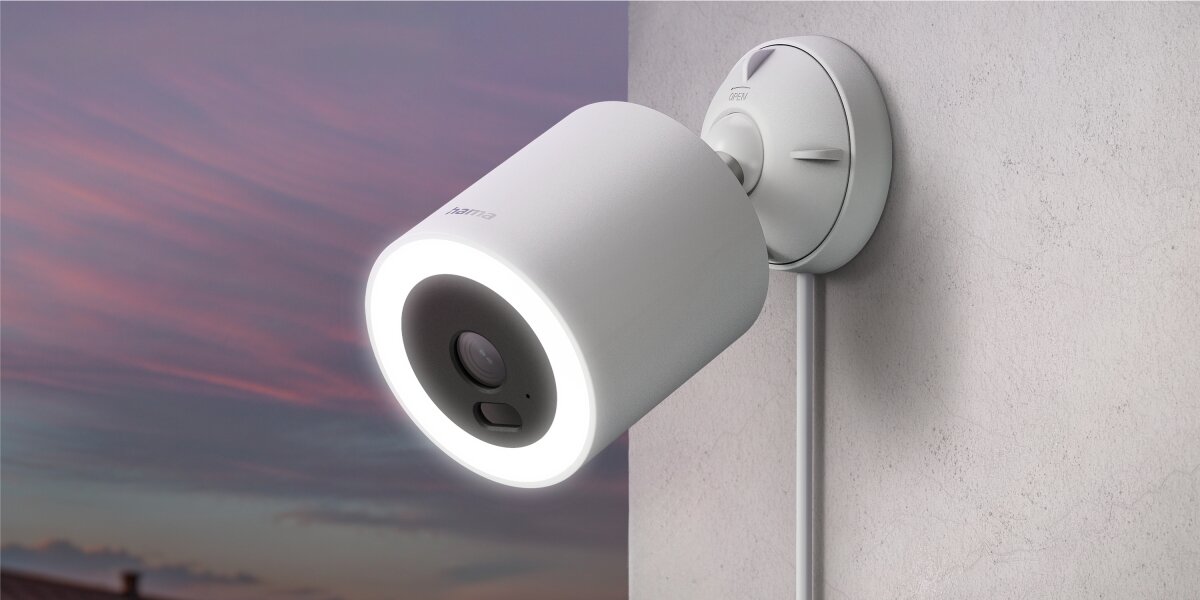 Kamera HAMA Smart Surveillance Zewnętrzna, Wi-Fi Biała zewnętrzna kamera Hama Smart Home z podświetlanym pierścieniem LED zamontowana na ścianie na tle wieczornego nieba.