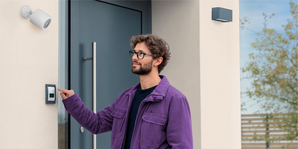 Kamera HAMA Smart Surveillance Zewnętrzna, Wi-Fi Mężczyzna dzwoniący do drzwi wejściowych monitorowanych przez inteligentną kamerę zewnętrzną Hama zamontowaną powyżej.