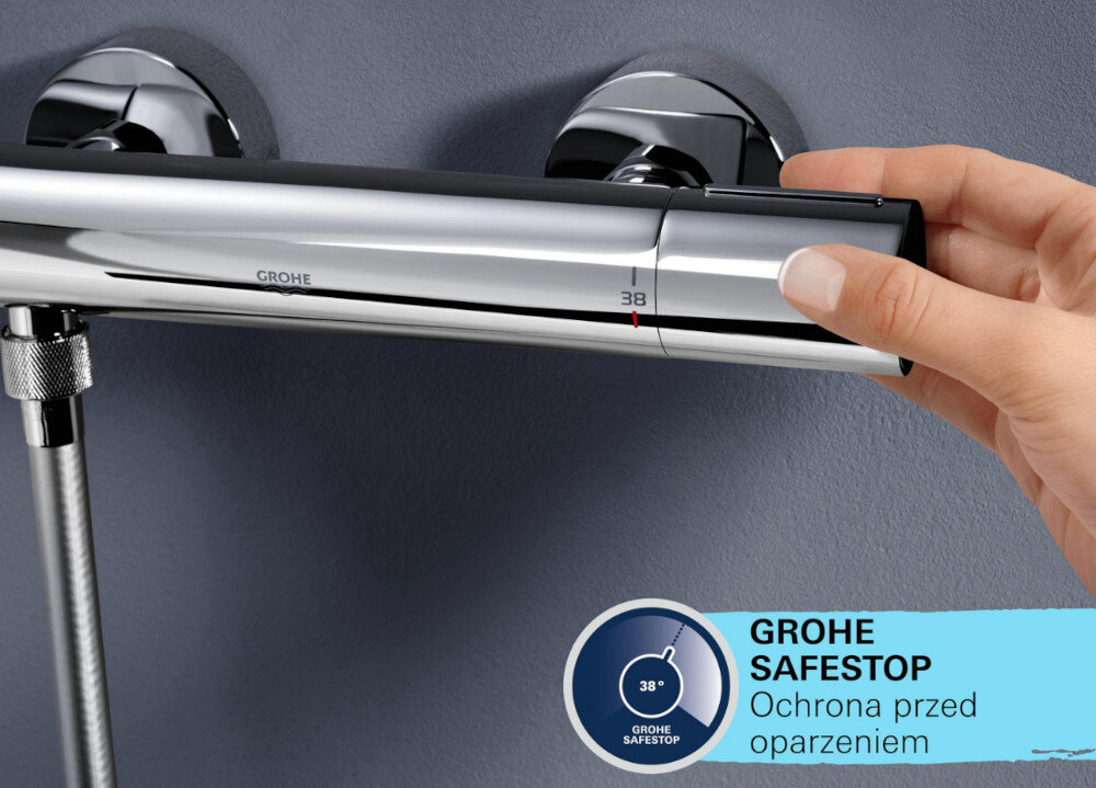 Osoba reguluje temperaturę na chromowanym panelu prysznicowym z widocznym logo GROHE i oznaczeniem 38 stopni. Na dole znajduje się napis \'GROHE SAFESTOP Ochrona przed oparzeniem\'.
