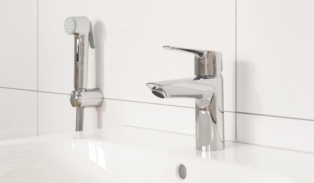 BATERIA UMYWALKOWA GROHE Start S S 23123003 CHROM Na zdjęciu widoczna jest chromowana bateria umywalkowa zamontowana na białej umywalce, obok uchwytu z rączką prysznicową. Całość utrzymana jest w jasnej, minimalistycznej aranżacji łazienki