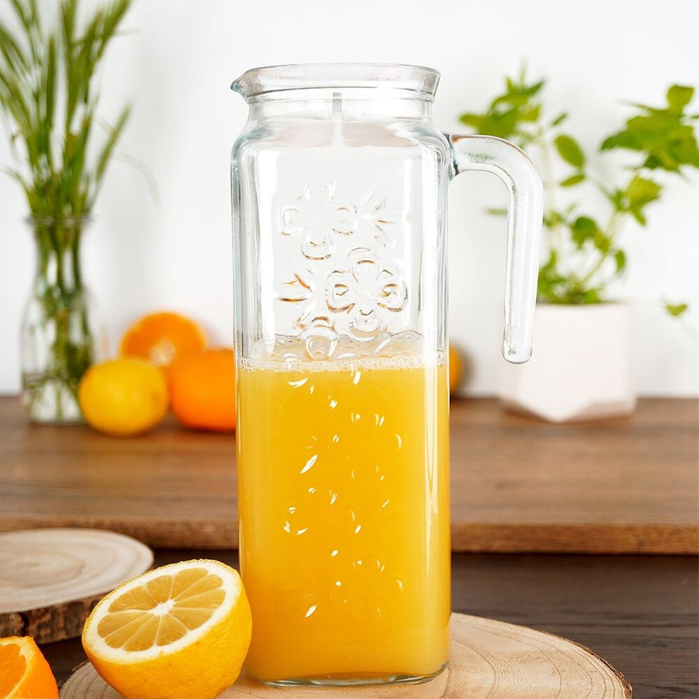 Szklany dzbanek z pokrywką o pojemności 1,2 l, wypełniony pomarańczowym napojem, stoi na drewnianym blacie obok przekrojonej cytryny. W tle widoczne są pomarańcze i zielone rośliny.