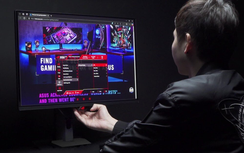 Monitor ASUS ROG Swift PG27AQWP-W Osoba siedząca tyłem do widza i sięgająca dłonią pod dolną ramkę monitora. Na ekranie wyświetlone jest czerwone menu ustawień na tle pokoju gracza