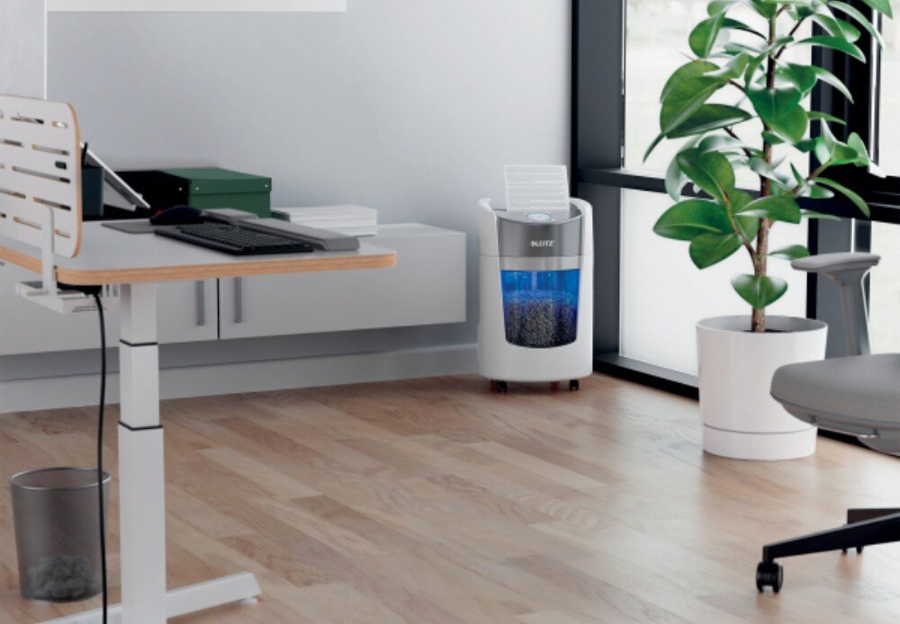 W biurze znajduje się niszczarka z widocznym logo \'LEITZ\', obok której stoi roślina w doniczce. Na biurku leży klawiatura i myszka, a obok znajduje się zamknięty laptop.