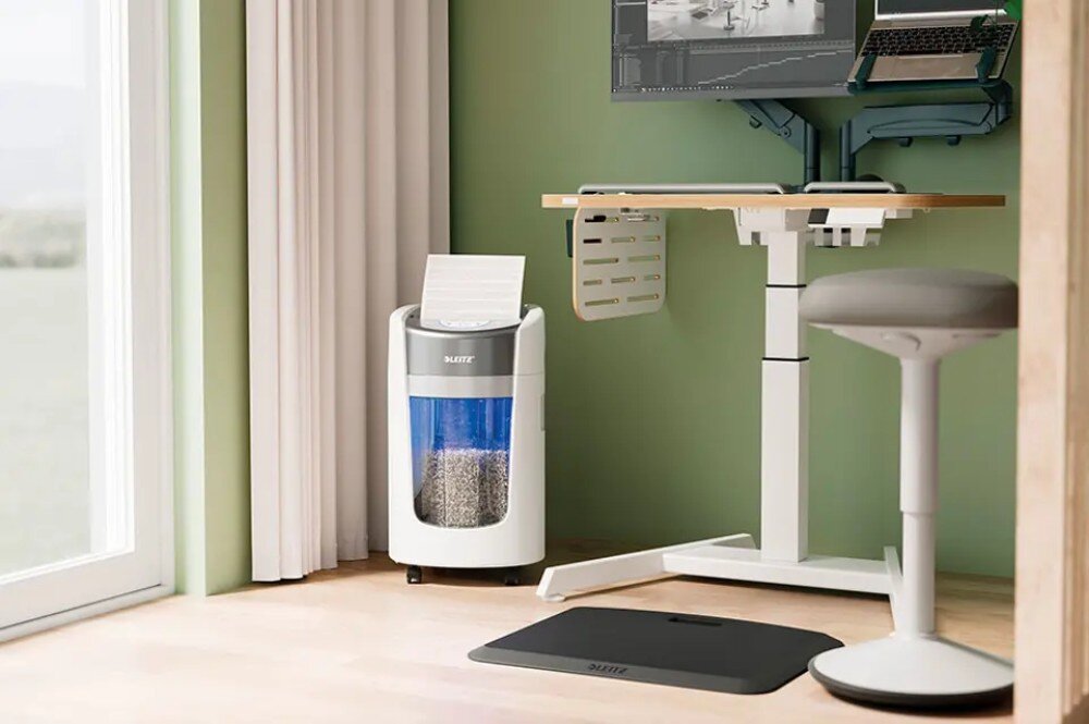 Niszczarka LEITZ IQ OptiMax Home Office 225 P5 Półzbliżenie na białą niszczarkę na kołkach, stojącą na jasnej, drewnianej podłodze, z niebieskim, przezroczystym zbiornikiem wypełnionym pociętym papierem i arkuszami papieru czekającymi na zniszczenie, obok białego biurka ze regulacją wysokości i szarego, stołka