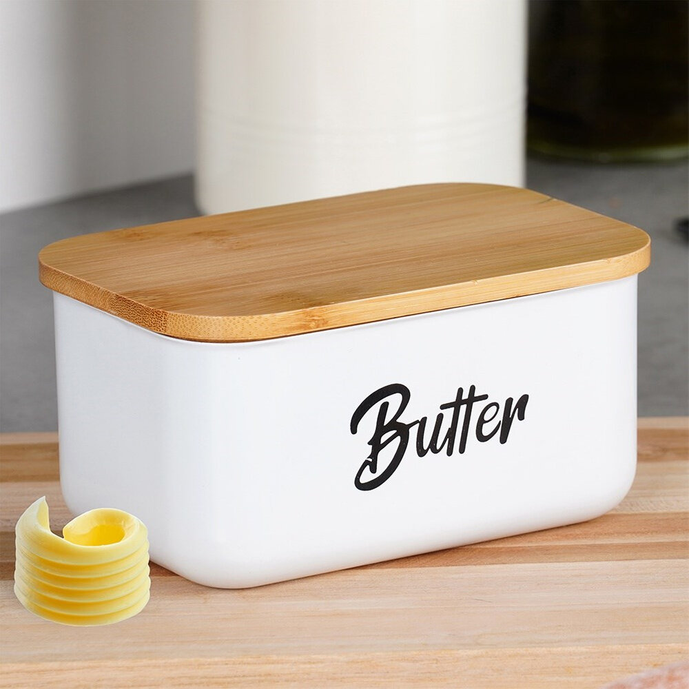 Biała maselniczka z bambusową pokrywką, na której widnieje napis \'Butter\'. Obok znajduje się kilka zrolowanych kawałków masła.
