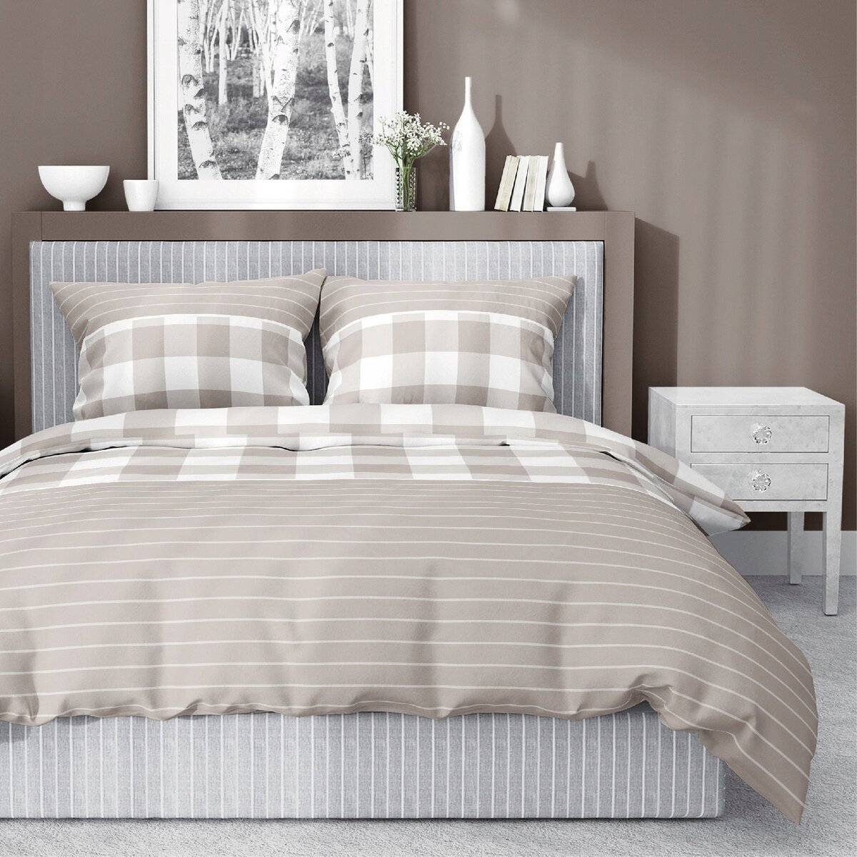 Na łóżku znajduje się komplet pościeli w odcieniach beżu i bieli z wzorem w kratkę i paski. W tle widoczna jest szafka nocna oraz dekoracje na półce, w tym obraz z motywem drzew.