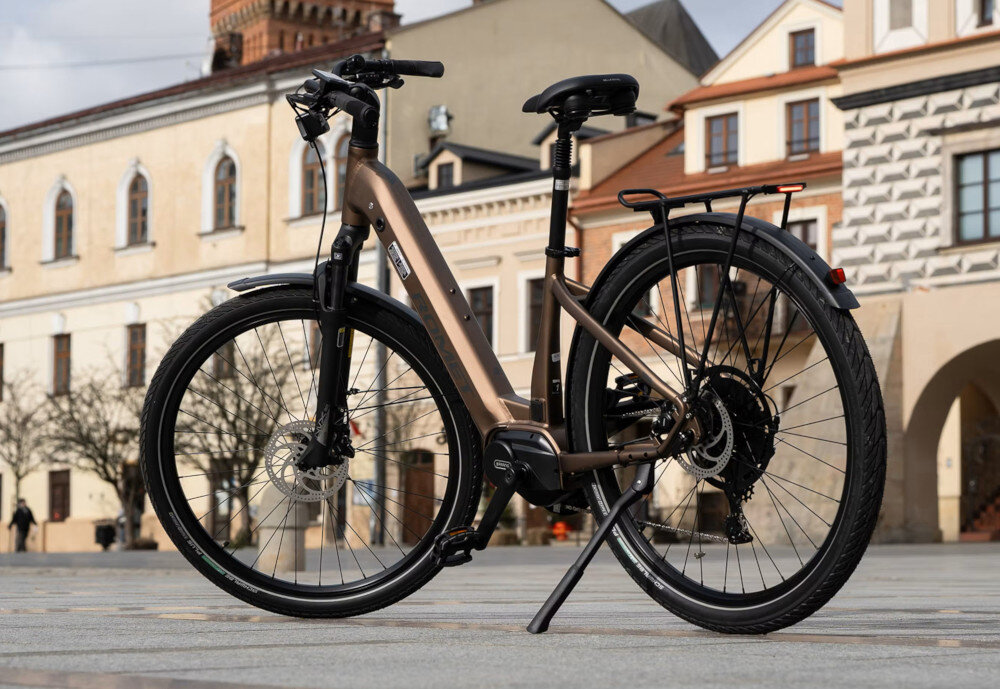 Rower elektryczny ROMET E-Modeco URB 2.0 U17 28 cali Brązowo-czarny miasto deptak rower oparty o stopkę widoczny od tyłu pod skosem uniwersalny rower elektryczny środek codziennego transportu opcja dosłownie dla każdego wymaga on również pracy mięśni nóg elektryczny układ napędowy działa tu niejako równolegle