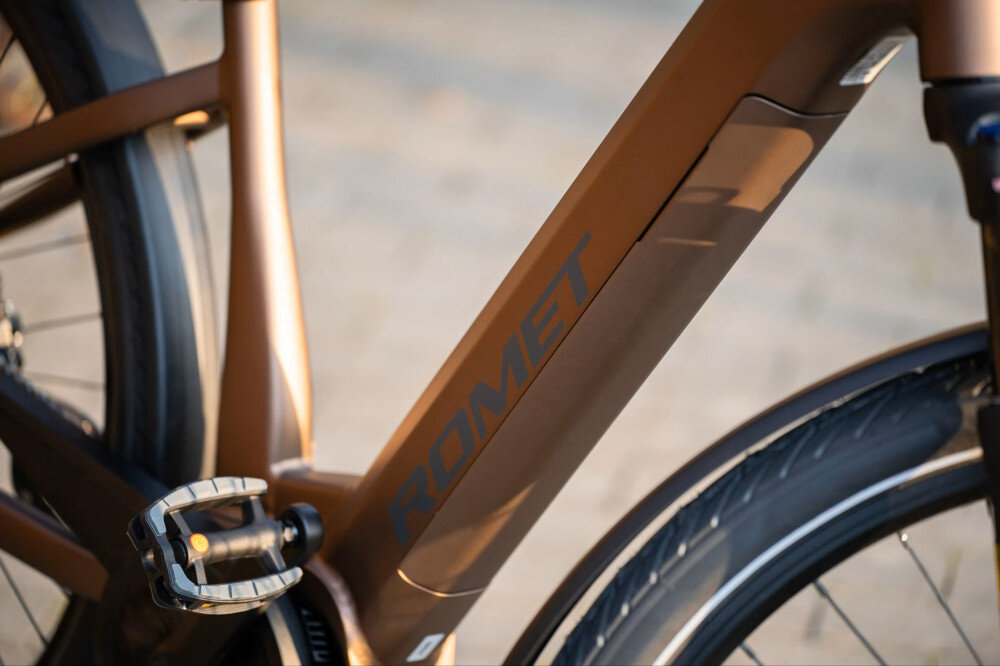 Rower elektryczny ROMET E-Modeco URB 2.0 U17 28 cali Brązowo-czarny zbliżenie na baterię rozmazane tło zasięg do 100 km bateria Li-Ion o napięciu 36 V i pojemności 15 Ah (540 Wh) bateria waży około 4 kg czas ładowania około 4-8 godzin żywotność około 500 ładowań bateria w ramie w zestawie dwa kluczyki zintegrowana z konstrukcją roweru