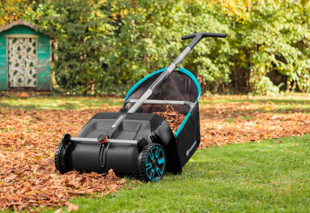 Na trawniku znajduje się czarny zbieracz do liści z niebieskimi akcentami i logo \'GARDENA\'. W tle widoczna jest zielona altana oraz drzewa z jesiennymi liśćmi.