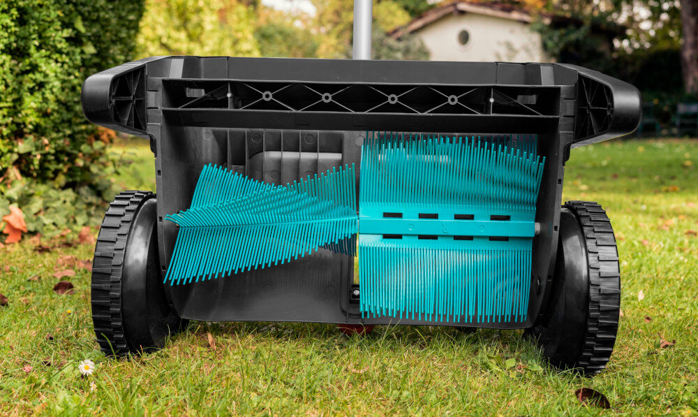 Na obrazie widoczny jest zbieracz do liści z niebieskimi szczotkami i czarną obudową, umieszczony na trawie. W tle znajduje się ogród z zielenią i budynkiem.