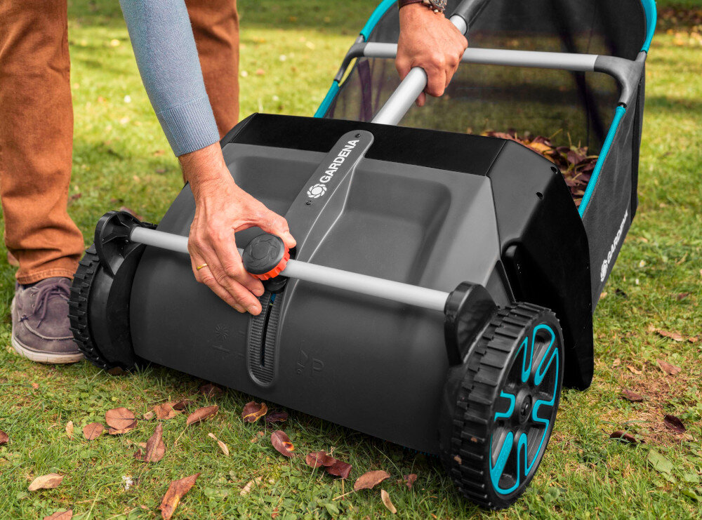 Osoba używa zbieracza do liści marki GARDENA na trawniku. Urządzenie ma duże koła i uchwyt z widocznym logo GARDENA.
