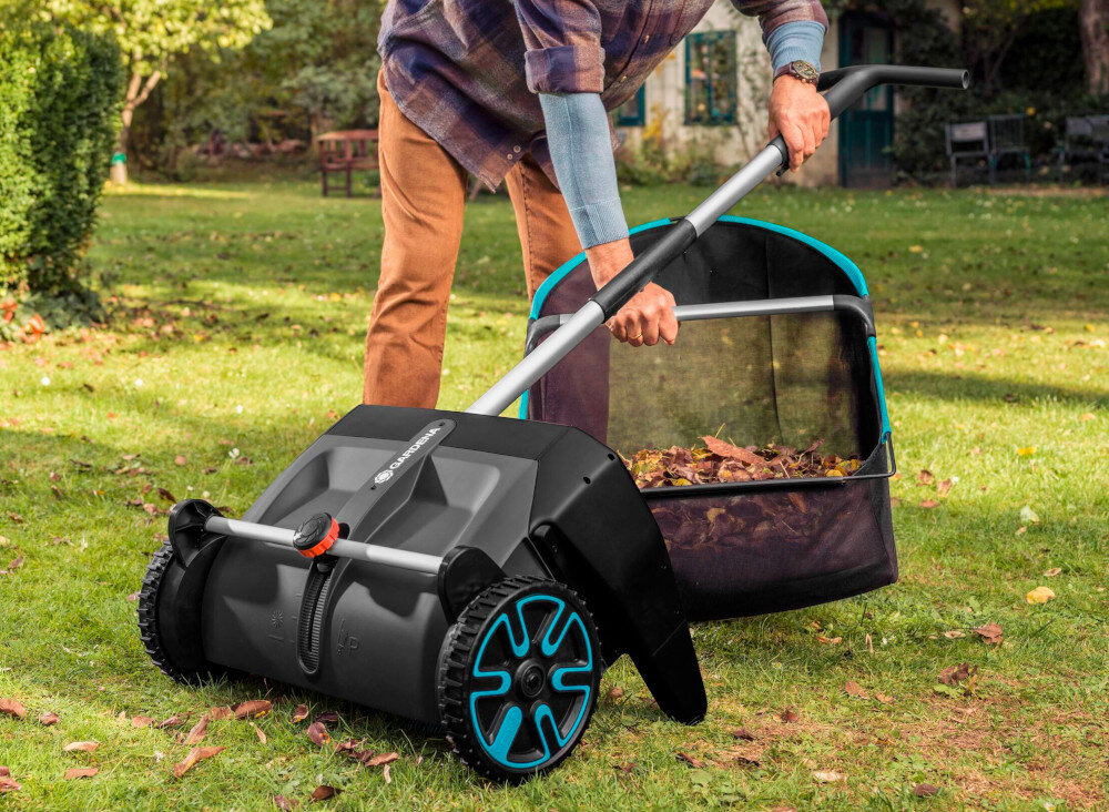 Osoba używa zbieracza do liści na trawniku, w tle widoczne są drzewa i budynek. Na urządzeniu znajduje się logo \'GARDENA\'.