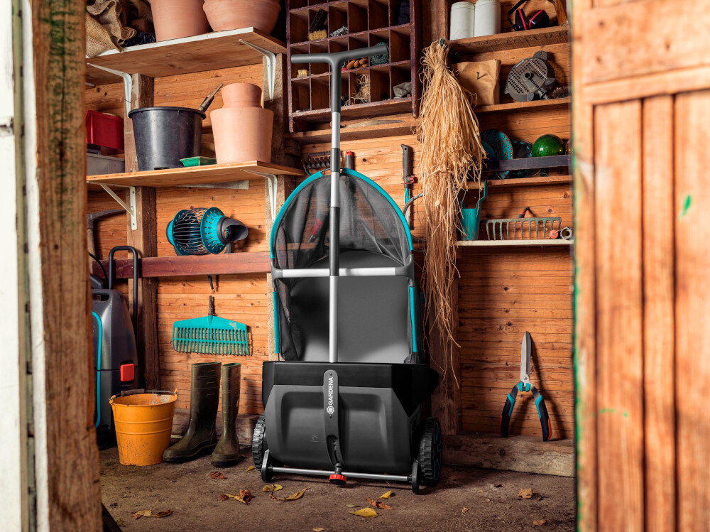 W drewnianym pomieszczeniu znajduje się zbieracz do liści z logo \'GARDENA\', otoczony narzędziami ogrodniczymi i doniczkami. Na podłodze leżą liście, a na półkach widać różne przedmioty ogrodnicze.