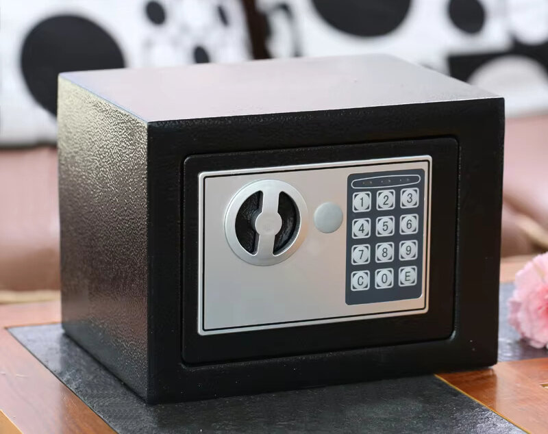 Czarny sejf z zamkiem elektronicznym i klawiaturą numeryczną z cyframi od 1 do 9 oraz przyciskami C, 0, E. Sejf stoi na drewnianej powierzchni, w tle widoczne są poduszki z czarno-białymi wzorami.