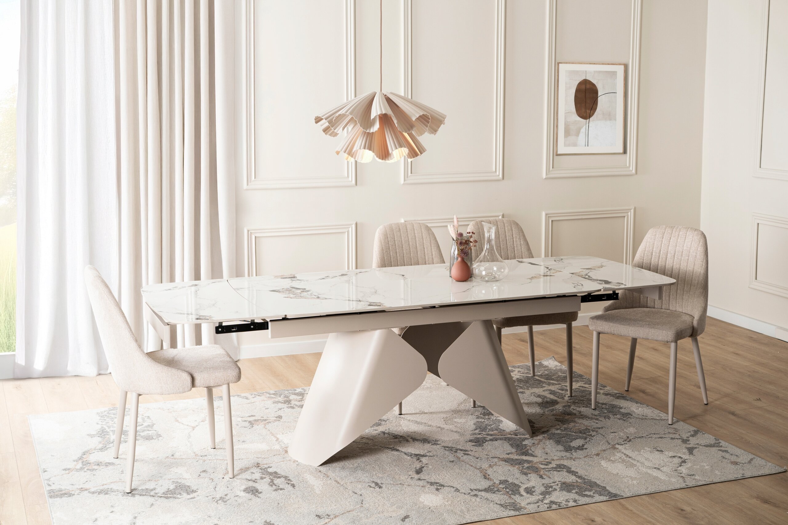 Na obrazie znajduje się elegancki, biały stół ceramiczny z rozkładanym blatem, otoczony czterema krzesłami. W tle widać jasne zasłony, ścianę z panelami oraz wiszącą lampę i obraz.