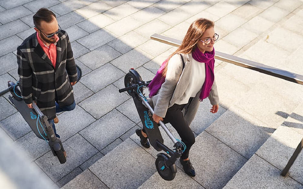 Hulajnoga elektryczna MOTUS Scooty 10 Lite Gen 4 30km 350W Amortyzacja 10 cali Szary widok z góry dwie osoby idą po chodniku w rękach trzymają złożone hulajnogi składana konstrukcja łatwy transport zwolnienie mechanizmu pociągnięciu zatrzasku popchnięcie kierownicy do przodu szybkie złożenie hulajnogi