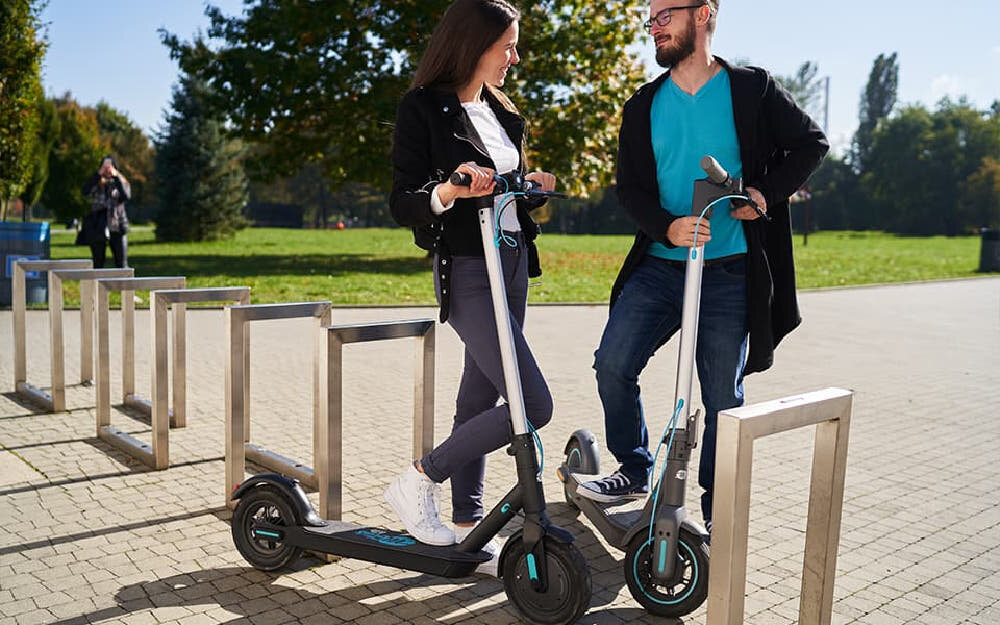 Hulajnoga elektryczna MOTUS Scooty 10 Lite Gen 4 30km 350W Amortyzacja 10 cali Szary dwie osoby stoją opierając się o hulajnogi hulajnogi postawione na stopkach w tle chodnik i drzewa składana stopka wytrzymała nóżka w centralnym punkcie ciężkości