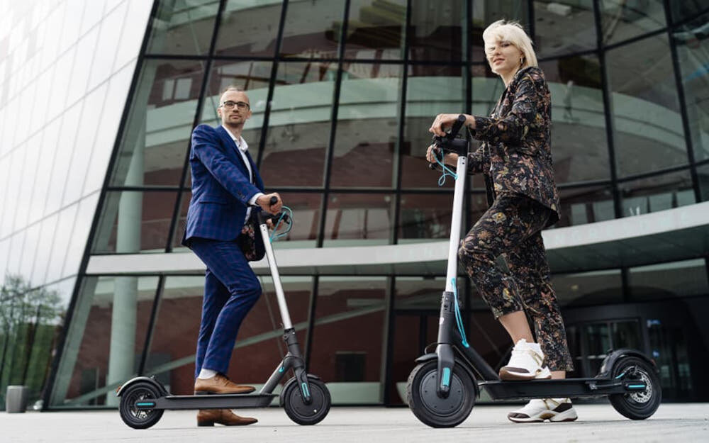 Hulajnoga elektryczna MOTUS Scooty 10 Lite Gen 4 30km 350W Amortyzacja 10 cali Szary dwie osoby stoją oparte o hulajnogi chodnik w tle budynek system KERS odzyskiwanie energii kinetycznej bateria będzie starczała na dłużej większy zasięg